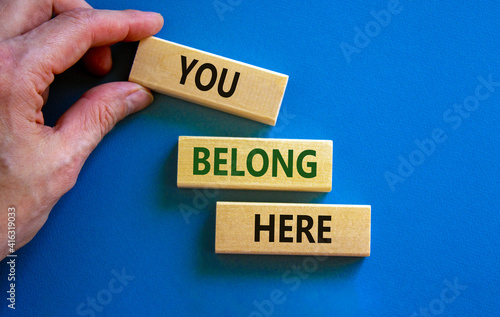 You belong here symbol. Wooden blocks with words 'You belong here' on beautiful blue background. Businessman hand. Diversity, business, inclusion and belonging concept. photo