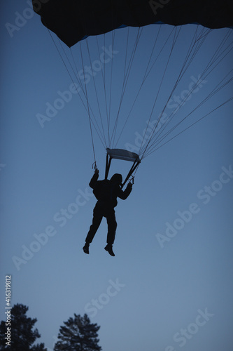 Dark silhouette of a basejumper under the canopy of a parachute, during a night jump, prepares for landing, flying over the crowns of trees, close-up.