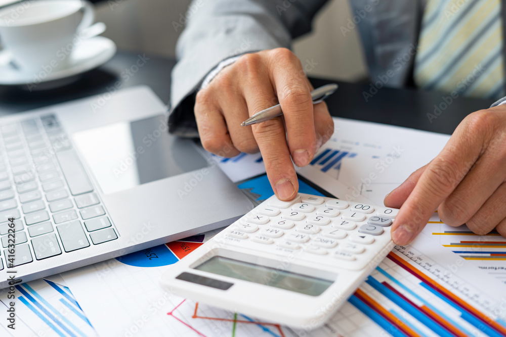 Asian male finance staff is calculating by using a calculator. Investment results to report to his boss at the meeting. On the table in the office, the concept of calculating investment results Asian male finance staff is calculating by using a calculator. Investment results to report to his boss at the meeting. On the table in the office, the concept of calculating investment results