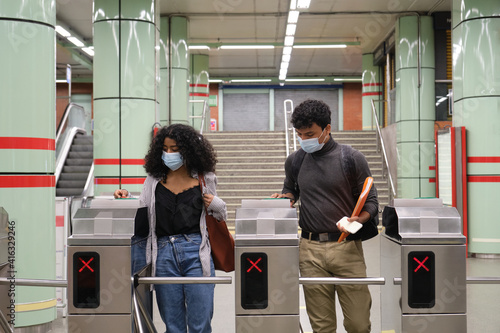 Young latin couple wearing protective face mask walking through subway turnstile at the train or metro station. New normal at public transport.