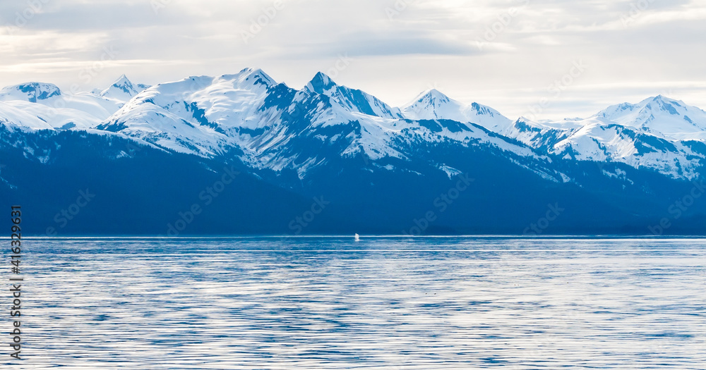 Snow-capped mountains along the coast of Alaska with the blow from a Humpback Whale surfacing visible in the distance