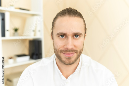 Handsome European man with light brown hair, gray eyes and beard looking at camera, smiling friendly, posing, confident, wearing white formal shirt, workplace, shelf with documents in the background