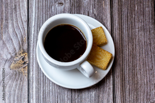 A Black coffee in a cup on a saucer with biscuits on the side laid on a wooden table
