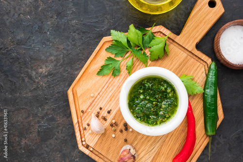 Chimichuri, Argentine green sauce for the steaks with fresh parsley, garlic, olive oil and oregano in a white mortar on a wooden board on a dark concrete background. Sauce recipes.