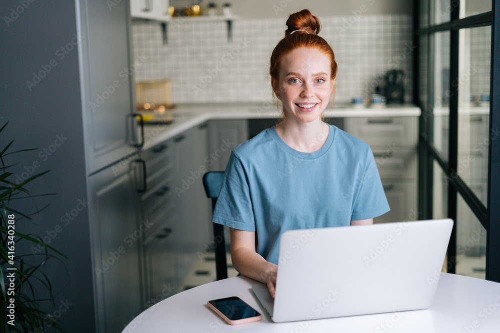 Front view of cheerful redhead young woman working typing on laptop computer sitting at table in kitchen room. Concept of leisure activity red-haired female at home during self-isolation.
