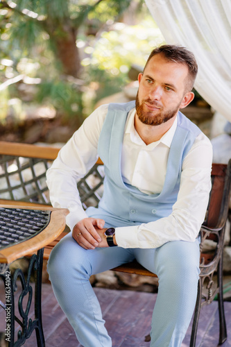 A groom with a beard in a blue suit sits on a chair in the park. 