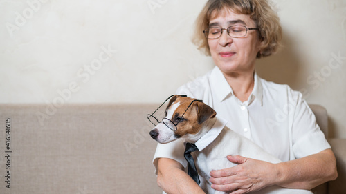 An elderly caucasian woman is holding a smart dog Jack Russell Terrier wearing glasses and a tie