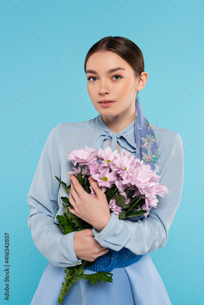 elegant young woman looking at camera while holding pink chrysanthemums isolated on blue elegant young woman looking at camera while holding pink chrysanthemums isolated on blue