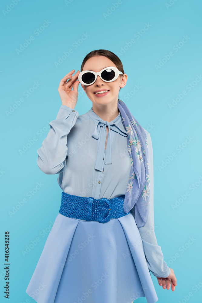 smiling fashionable woman touching sunglasses while posing isolated on blue smiling fashionable woman touching sunglasses while posing isolated on blue
