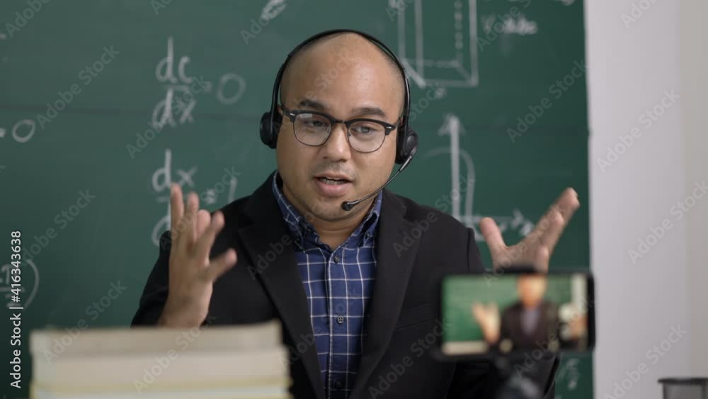 Indian young teacher man sitting wearing headset teaching online video conference live stream by smartphone. Asian teacher teaching mathematics class webinar online for students learning. Indian young teacher man sitting wearing headset teaching online video conference live stream by smartphone. Asian teacher teaching mathematics class webinar online for students learning.