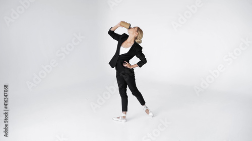 elegant ballerina in suit and pointe shoes drinking coffee to go while standing with hand on hip on white © LIGHTFIELD STUDIOS