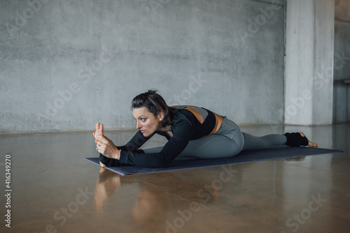 Caucasian beautiful woman practicing yoga at fitness center in hanumanasana, monkey king pose, in gray and black sportswear and barefoot. Indoor full length, side view. Healthy lifestyle.