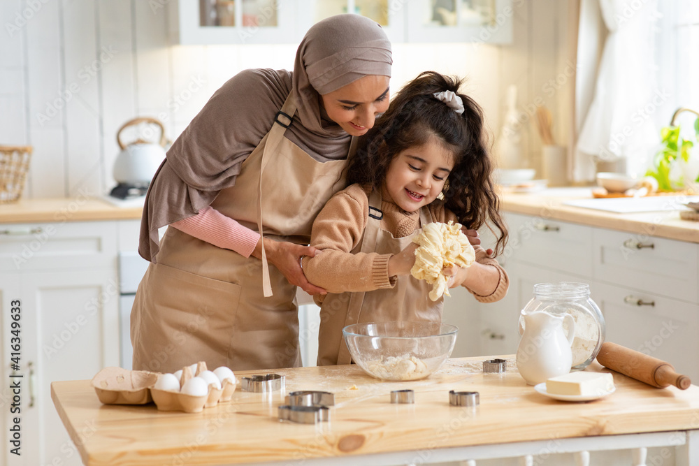 Cute little girl and her muslim mom kneading dough and having fun in kitchen
