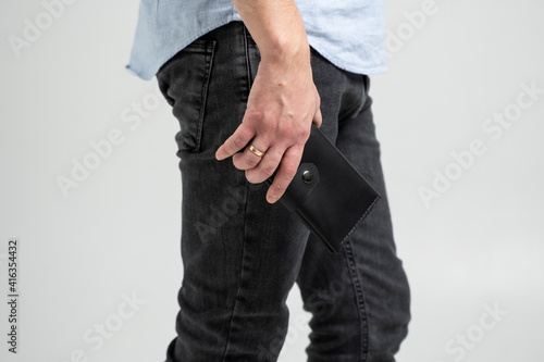Stylish handmade black leather wallet in a mens hand in blue shirt and black jeans on white background.