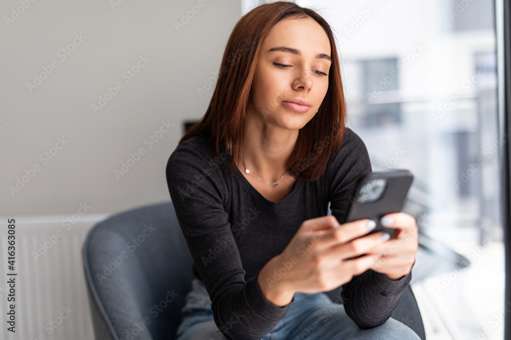 Sending message to him. Beautiful young smiling woman looking at her smart phone while sitting in a big comfortable chair at home Sending message to him. Beautiful young smiling woman looking at her smart phone while sitting in a big comfortable chair at home