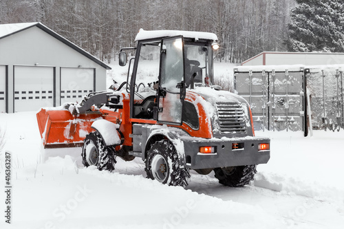 Little red tractor with snowplow removing snow during snowfall. Blizzard weather conditions. Winter time street maintenance.