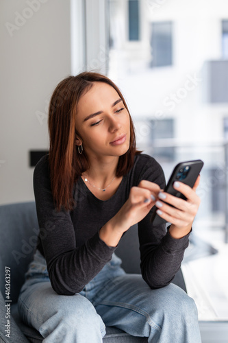 Smiling caucasian woman use smartphone sitting on armchair in bright living room.
