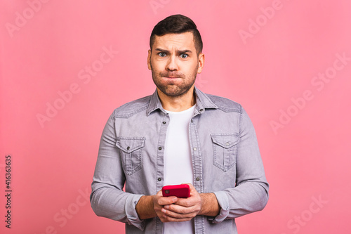 Portrait of a surprised amazed shocked casual young man looking at mobile phone isolated over pink background. Using phone, texting message. © denis_vermenko