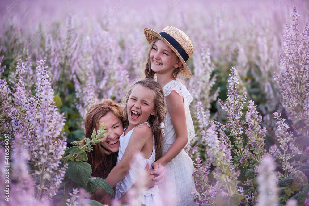 Young mother embraces two daughters among purple blooming sage field. Girls hug together, laugh, have fun. Dressed in white with straw hats. Traveling with children, maternal care. Close up portrait Young mother embraces two daughters among purple blooming sage field. Girls hug together, laugh, have fun. Dressed in white with straw hats. Traveling with children, maternal care. Close up portrait