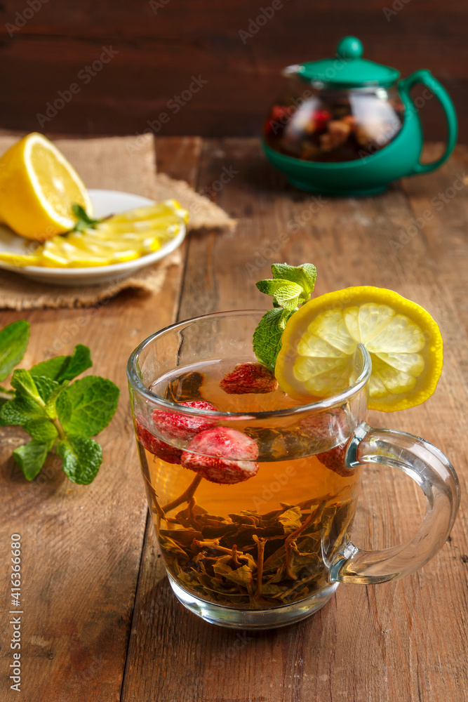 green tea in a glass cup with strawberries mint and lemon on a wooden table and a teapot and lemon in a plate and mint leaves. green tea in a glass cup with strawberries mint and lemon on a wooden table and a teapot and lemon in a plate and mint leaves.