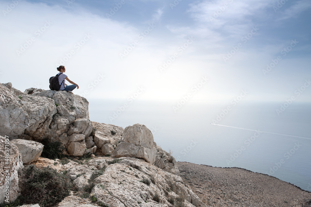 Young woman traveler with a backpack sits on rocks overlooking the sea and sky. Cyprus.