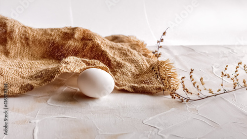 A white chicken egg, burlap and a stalk of dry grass on an isolated light background. The Concept of naturalness and Healthy Nutrition.