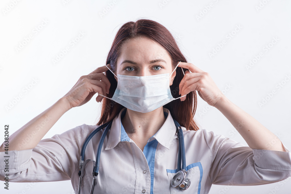 Portrait of a female doctor in a white uniform who puts on a medical mask in the studio on a white background
