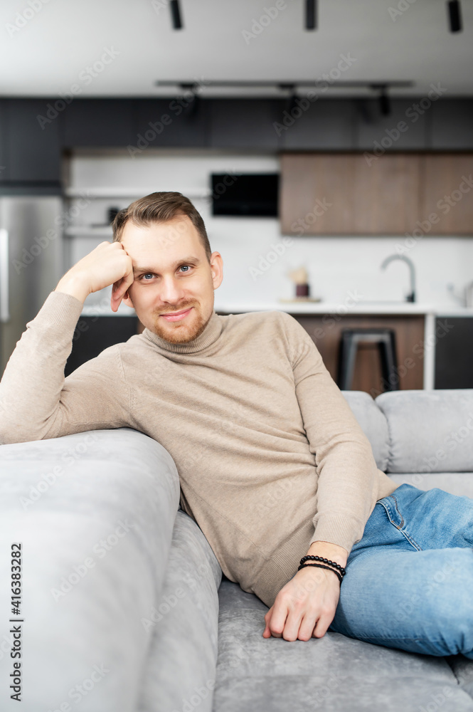 Confident and slender man with light brown hair, grey eyes and short neat bristle, sitting, head resting on one hand and looking at camera, flirting, friendly, posing, wearing beige sweater and jeans