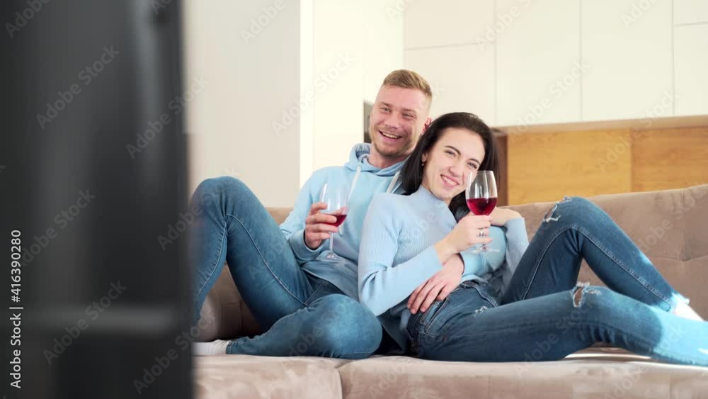 young cheerful couple watching funny movie on tv at home. A man and a woman or a boyfriend and a girlfriend are sitting on the couch with a glass of wine watching a TV series and laughing. young cheerful couple watching funny movie on tv at home. A man and a woman or a boyfriend and a girlfriend are sitting on the couch with a glass of wine watching a TV series and laughing.