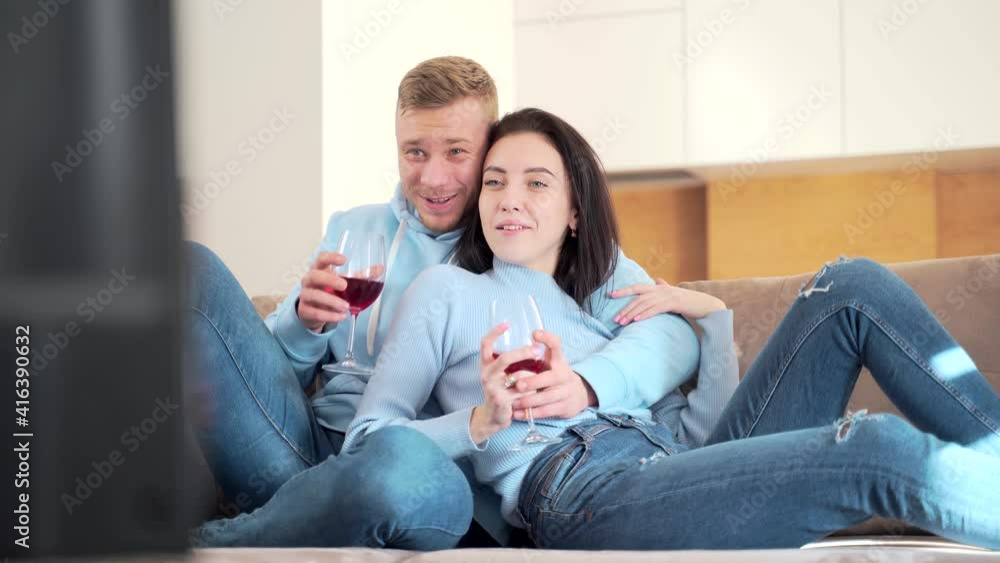 Cheerful young couple, boyfriend and girlfriend watching TV sitting on sofa and drinking wine. Family man and woman spending leisure time together enjoying watching television at home in new apartment Cheerful young couple, boyfriend and girlfriend watching TV sitting on sofa and drinking wine. Family man and woman spending leisure time together enjoying watching television at home in new apartment
