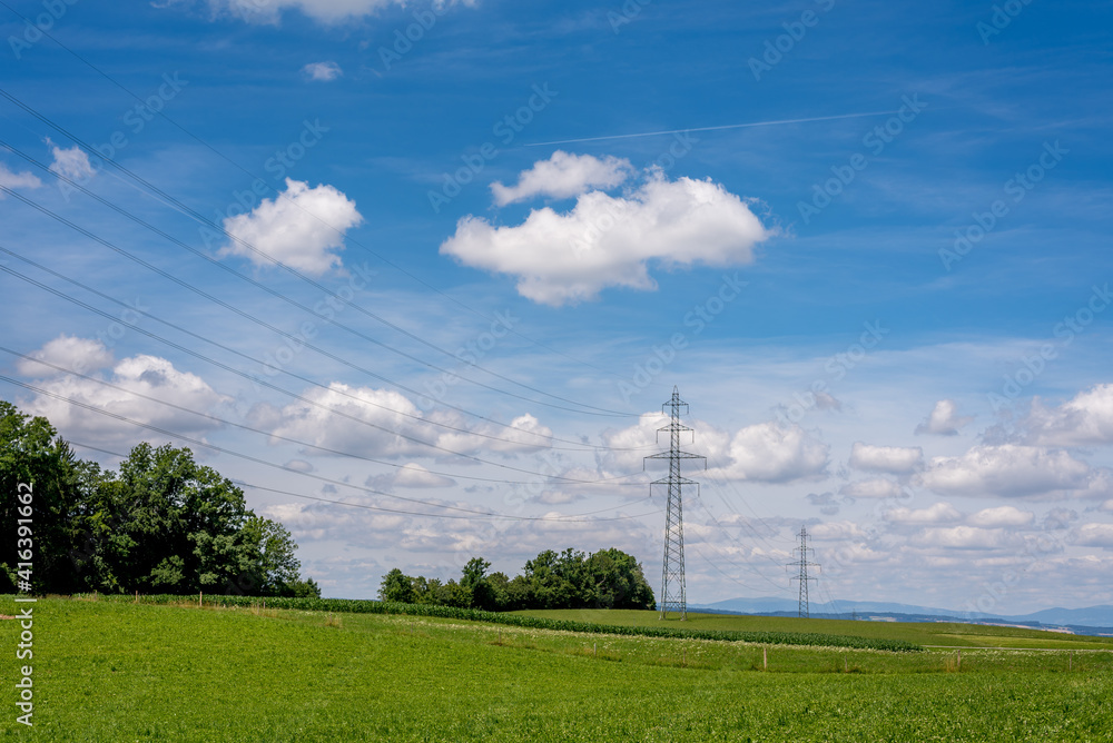 Obraz premium High voltage pylons and wires on the field. Landscape scene with agricultural field, trees, sky and clouds. Vevey, Switzerland.