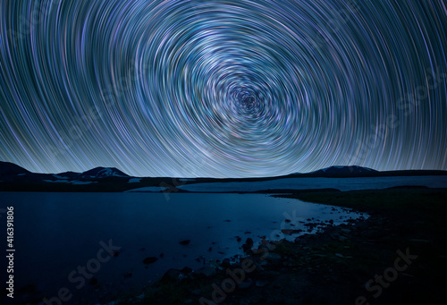 Beautiful small lake and mountains under the colorful star trails on the sky. Night time lapse photography.