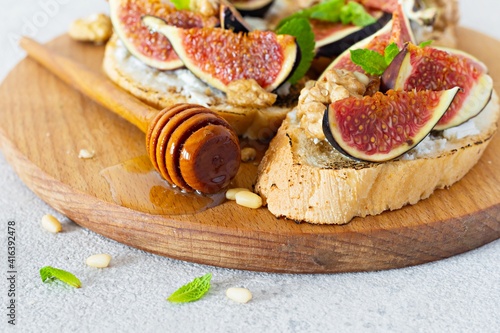Crispy bruschetta with soft ricotta, ripe figs, walnuts and pine nuts, mint and honey on a light background. Figs fruit toast on a wooden board with honey and walnut.
