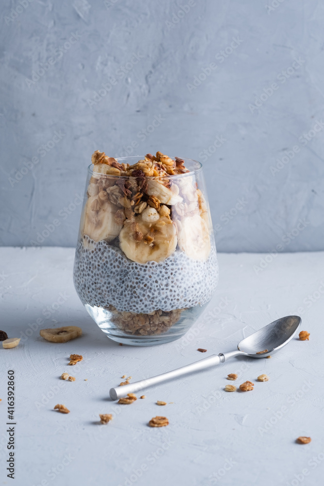 Healthy breakfast in a glass with banana, walnut, chio seeds, yogurt and muesli on a gray background with copy space.