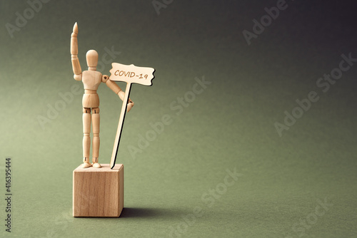 Coronavirus concept - wooden figure standing on a pedestal, raising hand and holding a sign with insctiption COVID-19 photo