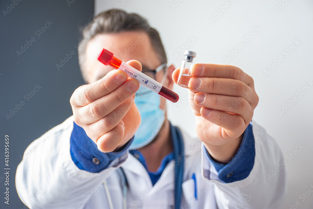Doctor, microbiologist or scientist holding in one hand laboratory test tube with blood, COVID-19 labeled and marked by positive result of analysis, in others - transparent vial coronavirus vaccine Doctor, microbiologist or scientist holding in one hand laboratory test tube with blood, COVID-19 labeled and marked by positive result of analysis, in others - transparent vial coronavirus vaccine