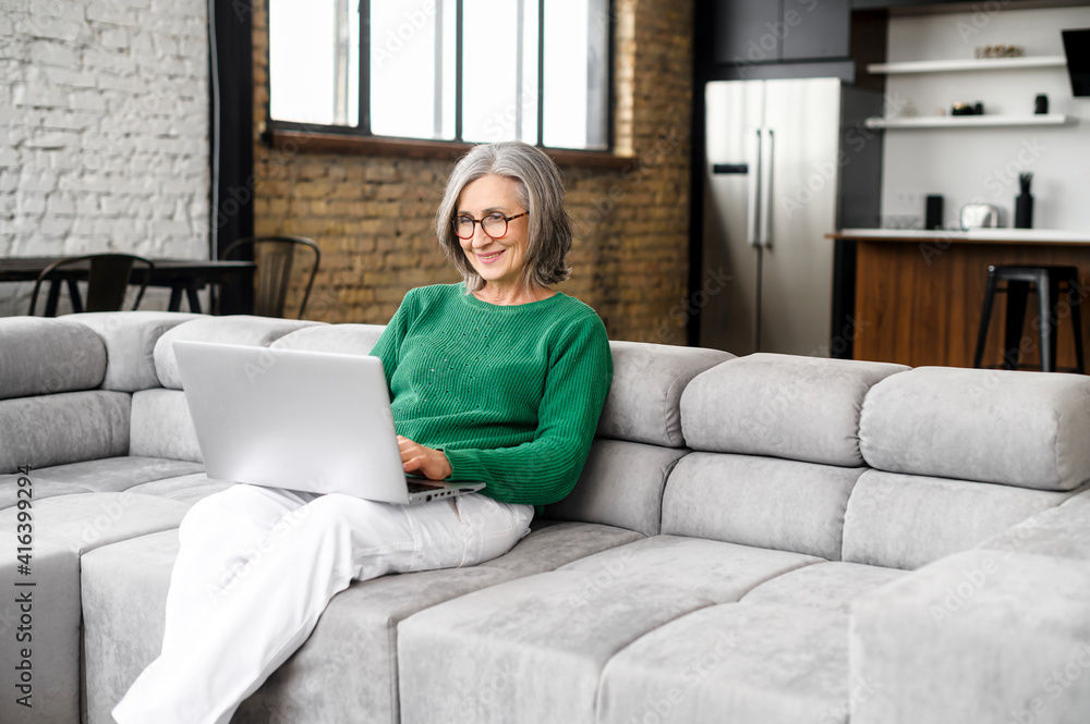 Charming senior elderly woman spends leisure time in networks on the computer, texting messages, watching movies. The mature lady works remotely with a laptop sitting on the sofa in cozy living room Charming senior elderly woman spends leisure time in networks on the computer, texting messages, watching movies. The mature lady works remotely with a laptop sitting on the sofa in cozy living room