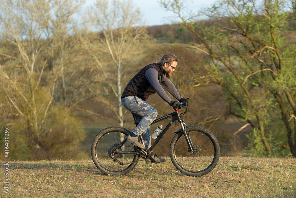 Sports brutal bearded guy on a modern mountain bike. Cyclist on the green hills in the spring.