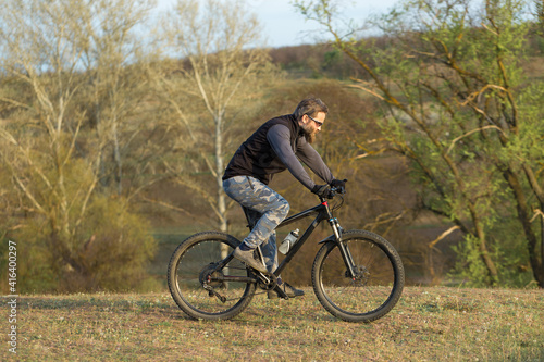 Sports brutal bearded guy on a modern mountain bike. Cyclist on the green hills in the spring.