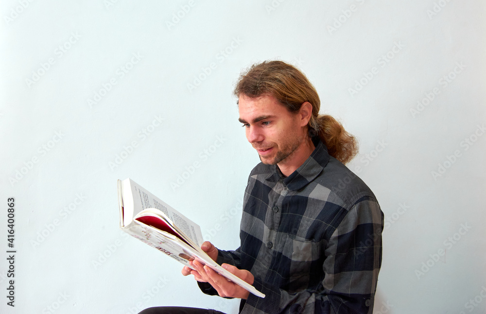 A handsome Caucasian man with long hair reading a book on the background of the white wall A handsome Caucasian man with long hair reading a book on the background of the white wall