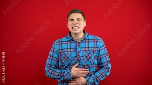 A young man has a stomach ache and he holds it in his hands. Shooting on a red background.