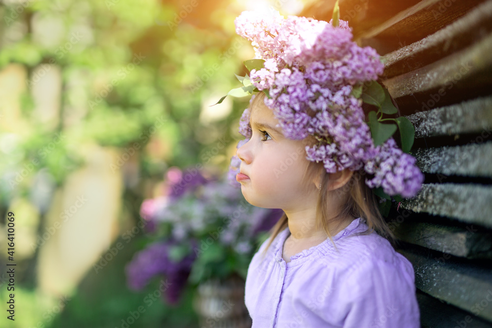 Obraz premium A girl with a wreath of lilac on her head looks away, pursing her lip. The pensive and offended look of a child. May evening sunset. The child looks to the side.