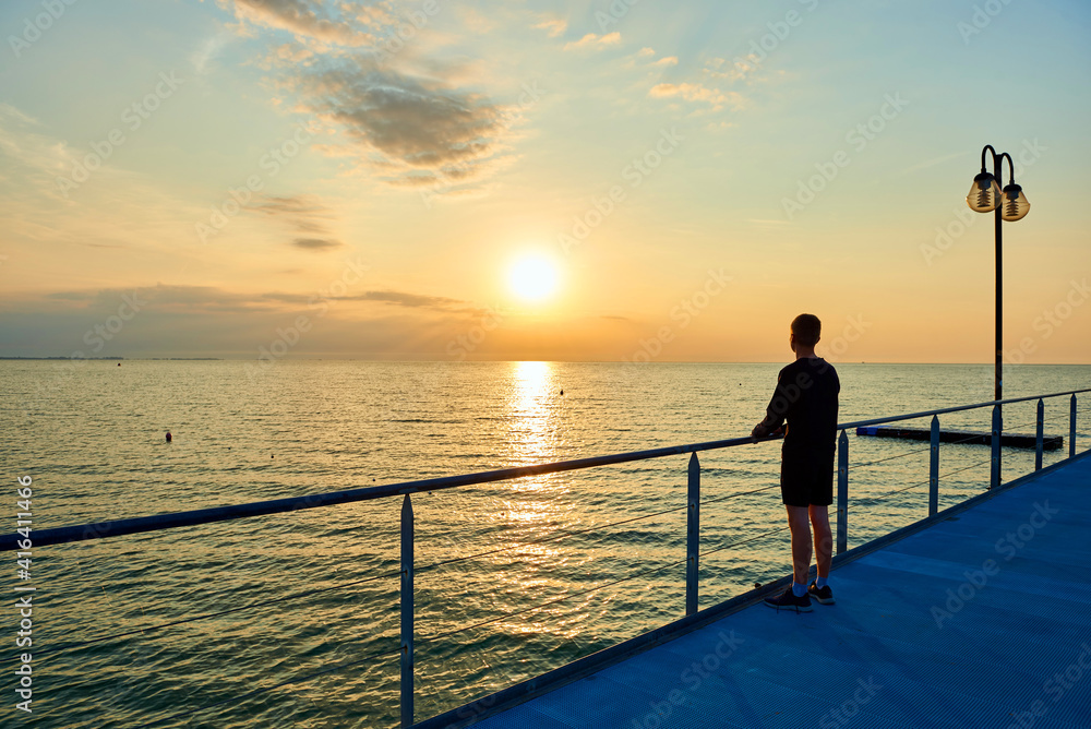 Fototapeta premium a boy is standing on a pier near Sea. Adriatic sea, sunset, waves and landscape. Lignano Sabbiadoro, Italy.
