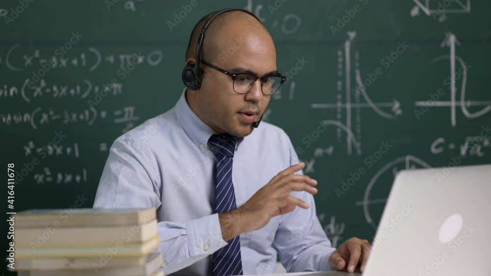 Indian young teacher man sitting wearing headset teaching online video conference live stream by laptop. Asian teacher teaching mathematics class webinar online for students learning. Indian young teacher man sitting wearing headset teaching online video conference live stream by laptop. Asian teacher teaching mathematics class webinar online for students learning.