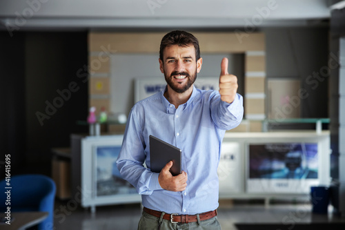 Smiling bearded businessman standing in lobby of export firm, holding tablet and showing thumbs up. Business is growing.