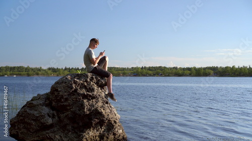 A young man sits on a stone by the river with a phone in his hands. The man is texting on his smartphone.