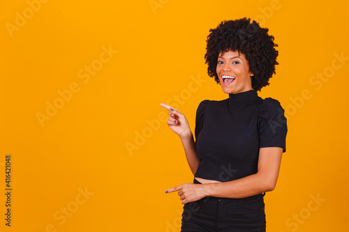 Horizontal shot of prettty young joyful African American female points with fore finger away, indicates copy space on blank wall, isolated over yellow background. Check this out. Ethnicity concept photo