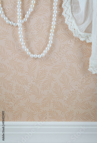 Draped lace curtain and strung pearls hang in front of a pink wallpaper background
