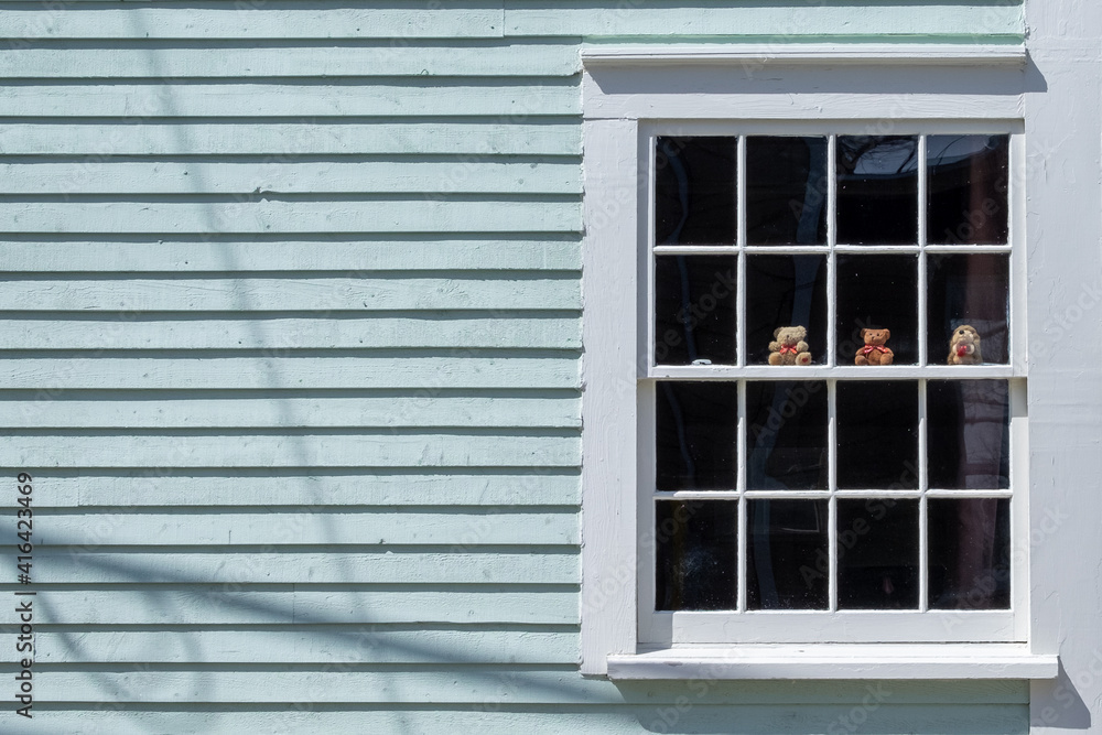 Fototapeta premium The exterior of a vintage mint green clapboard siding building with a multiple pane windows covered in white trim. There are two small teddy bears and a little toy beaver sitting on the window ledge. 