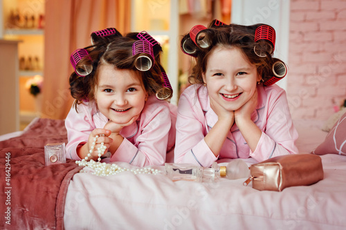 Little girls in pink pajamas do makeup while sitting in a room. Sisters with curlers play with makeup accessories in the children's room. The children are sitting on the bed with their mother's makeup